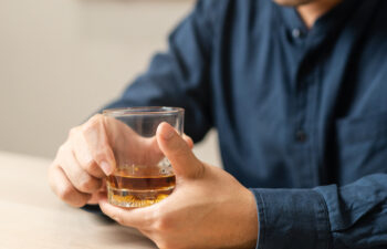 A person wearing a dark shirt sits at a table, holding a glass of amber-colored liquid, possibly whiskey or another alcoholic beverage.
