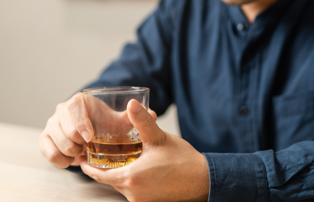 A person wearing a dark shirt sits at a table, holding a glass of amber-colored liquid, possibly whiskey or another alcoholic beverage.