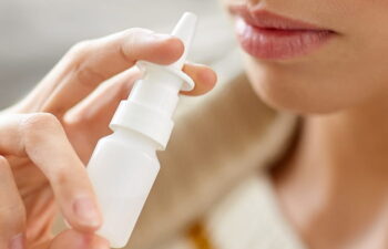A person holds a white nasal spray bottle close to their nose, preparing to use it.