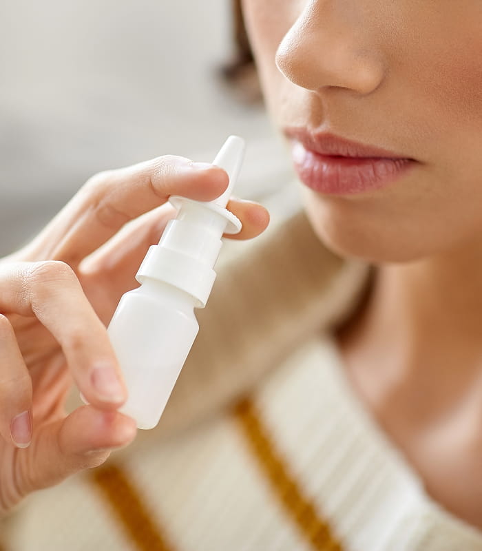 A person holds a white nasal spray bottle close to their nose, preparing to use it.