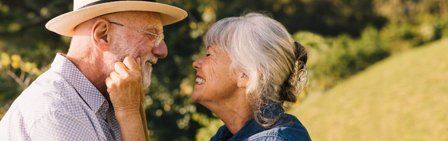 An older man wearing a hat and glasses smiles closely at an older woman outdoors; she is touching his face and smiling.
