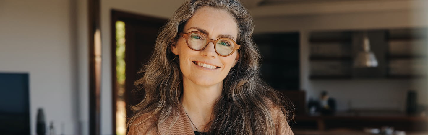 Woman with long gray hair and glasses smiling indoors, standing in a room with shelves and a hanging light in the background.