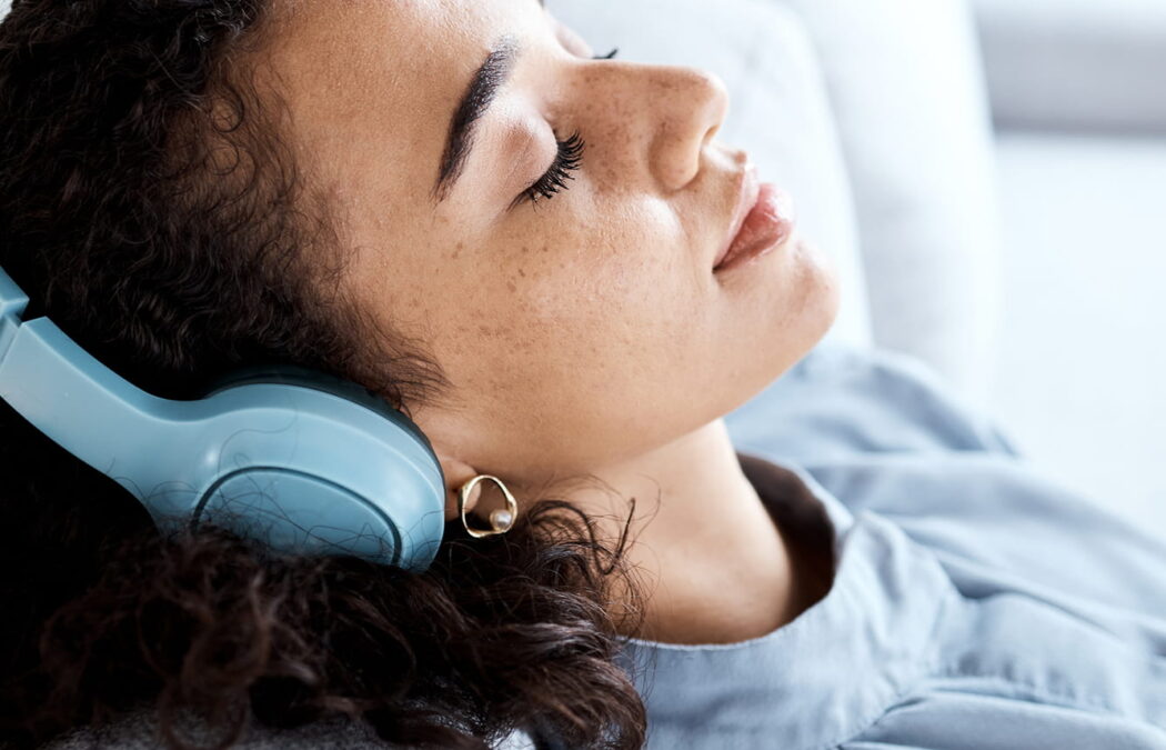 A person with curly hair wearing blue headphones is lying back on a couch with eyes closed, appearing relaxed.