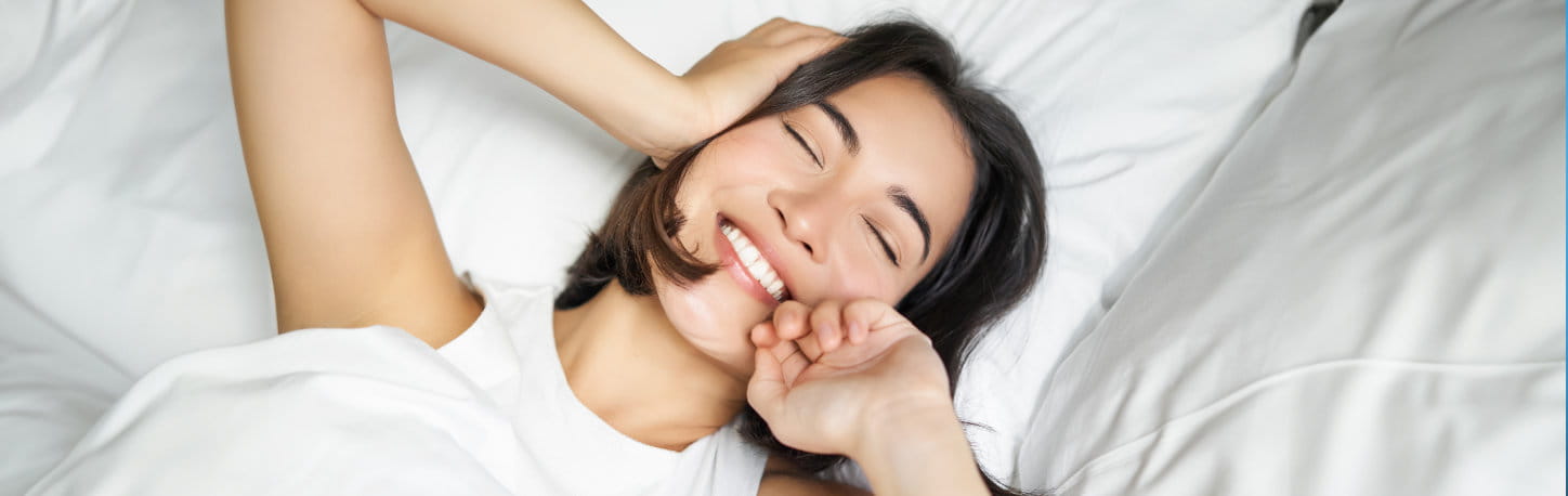 A woman lying in bed smiles with eyes closed, stretching her arms and appearing to wake up in the morning.