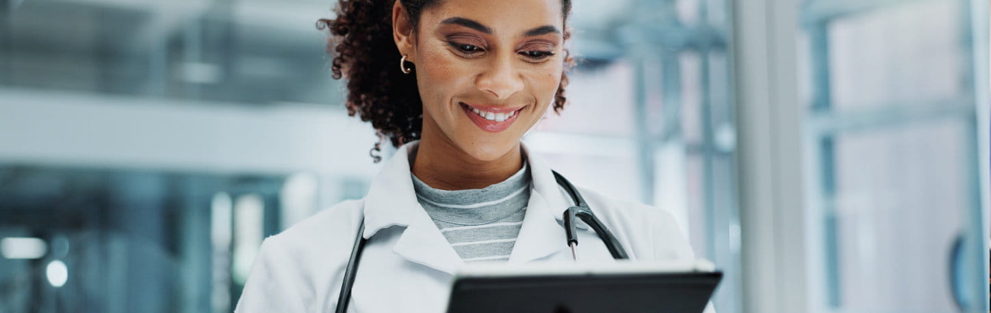 A doctor with a stethoscope around her neck looks at a digital tablet and smiles in a bright, modern medical office.