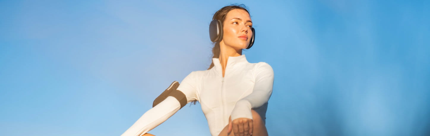 A woman in athletic wear and headphones stretches her arms outdoors against a clear blue sky.