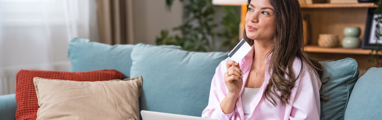 Woman sitting on a sofa holding a credit card and looking thoughtful, with a laptop on her lap and home decor in the background.
