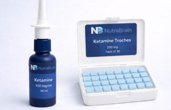 A bottle labeled "Ketamine 100 mg/mL" with a nasal spray top is next to an open case of "Ketamine Troches 200 mg," containing 30 light blue tablets.