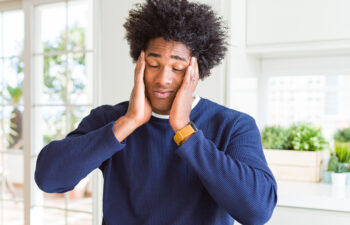 A person with closed eyes stands indoors, holding their head with both hands, appearing stressed or tired.
