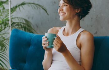 Side view young woman wears white tank shirt drink tea coffee sit on blue sofa couch stay at home hotel flat rest relax spend free spare time in living room indoors grey wall. People lounge concept