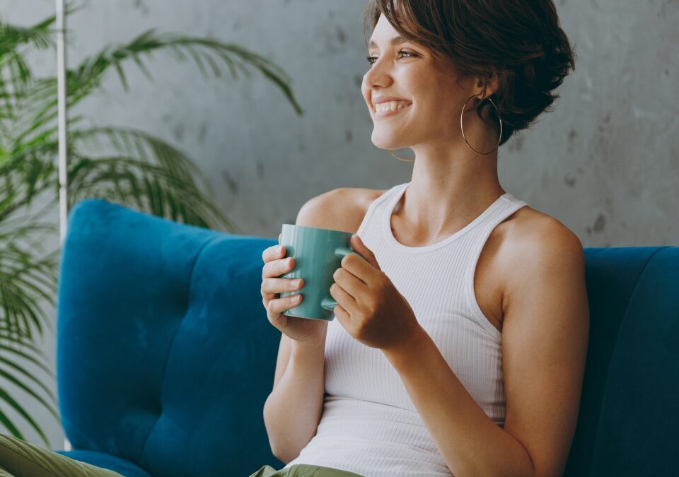 Side view young woman wears white tank shirt drink tea coffee sit on blue sofa couch stay at home hotel flat rest relax spend free spare time in living room indoors grey wall. People lounge concept