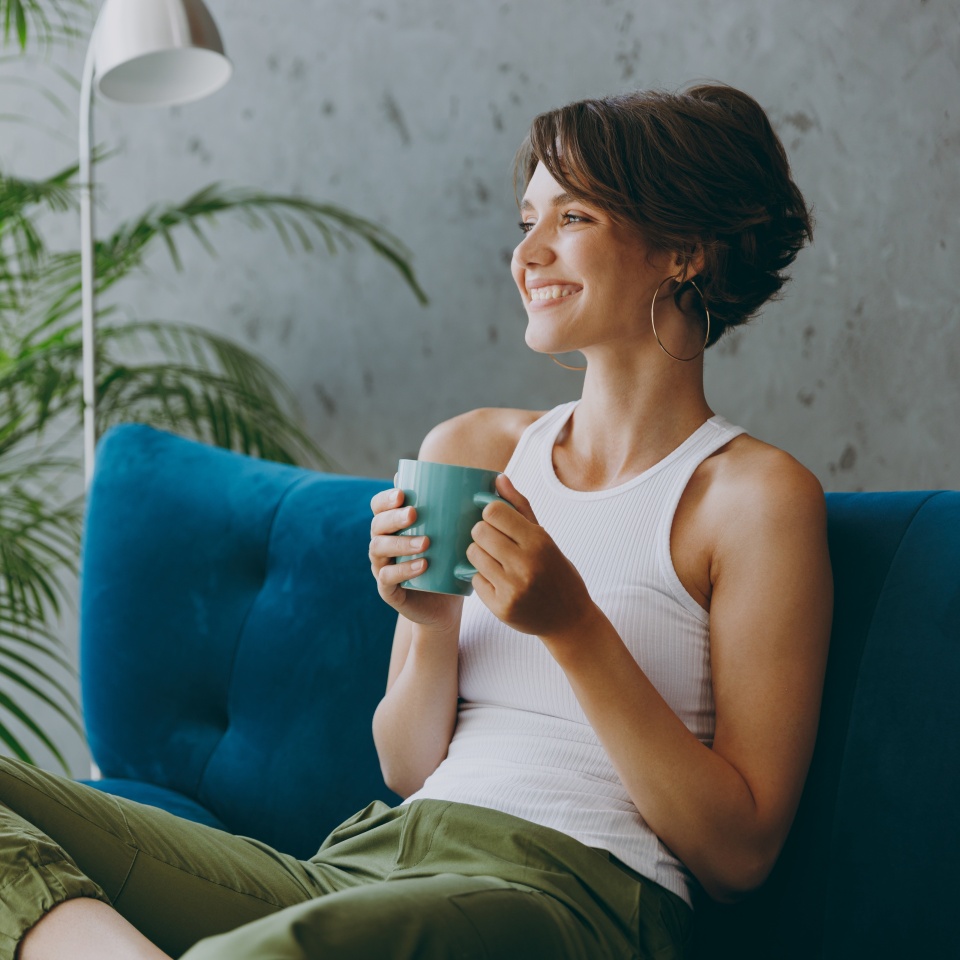 Side view young woman wears white tank shirt drink tea coffee sit on blue sofa couch stay at home hotel flat rest relax spend free spare time in living room indoors grey wall. People lounge concept