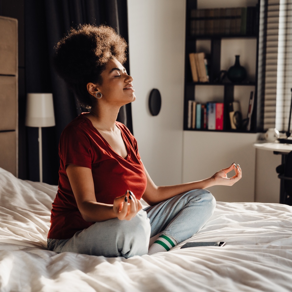 Smiling afro woman in casual wear meditating while sitting in lotus pose on bed at home