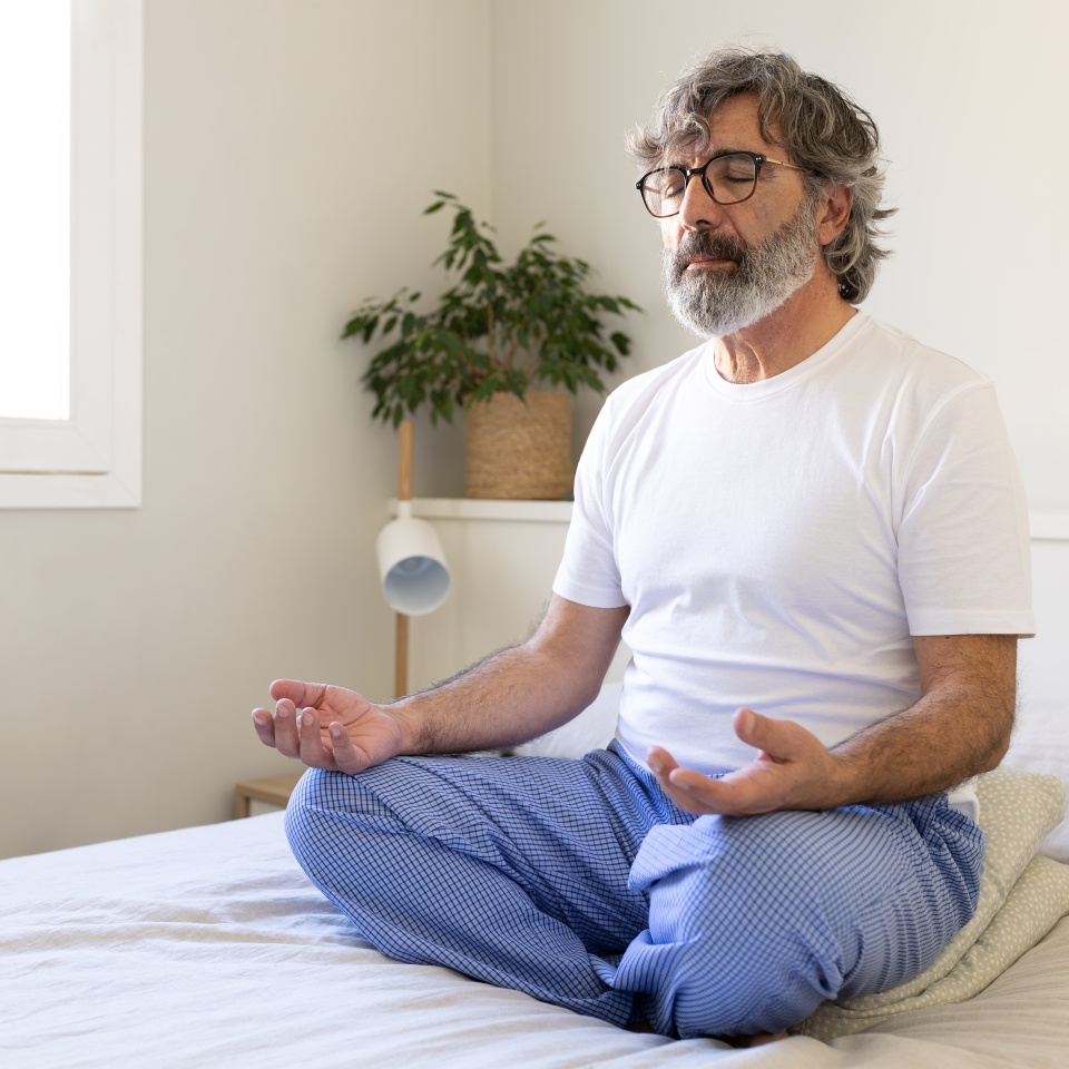 Mature adult man relaxing, doing meditation sitting on bed at home. Male meditating in the morning. Copy space. Wellness and mental health concept.