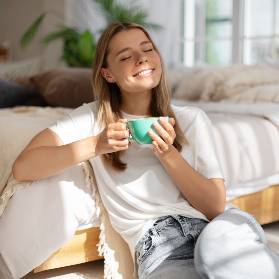 Young woman relaxes in sunlight, holding teal mug with eyes closed, enjoying a peaceful moment in a cozy bedroom. The room is bathed in natural light, featuring soft bedding, evoking calm and warmth