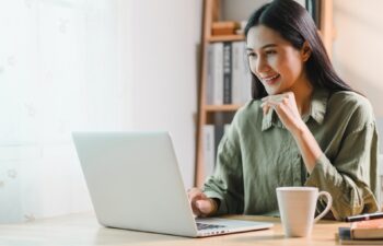 A woman working on laptop at wooden desk in bright home office, smiling while holding cup of coffee. room features bookshelves, lamp, and natural light from window