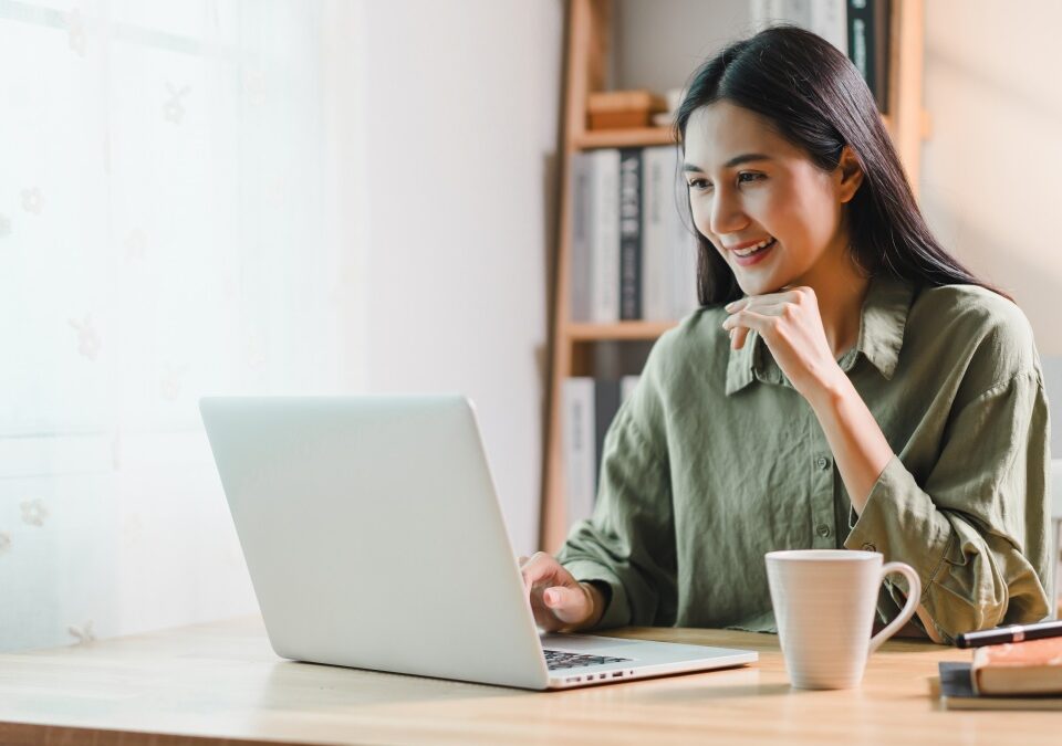 A woman working on laptop at wooden desk in bright home office, smiling while holding cup of coffee. room features bookshelves, lamp, and natural light from window