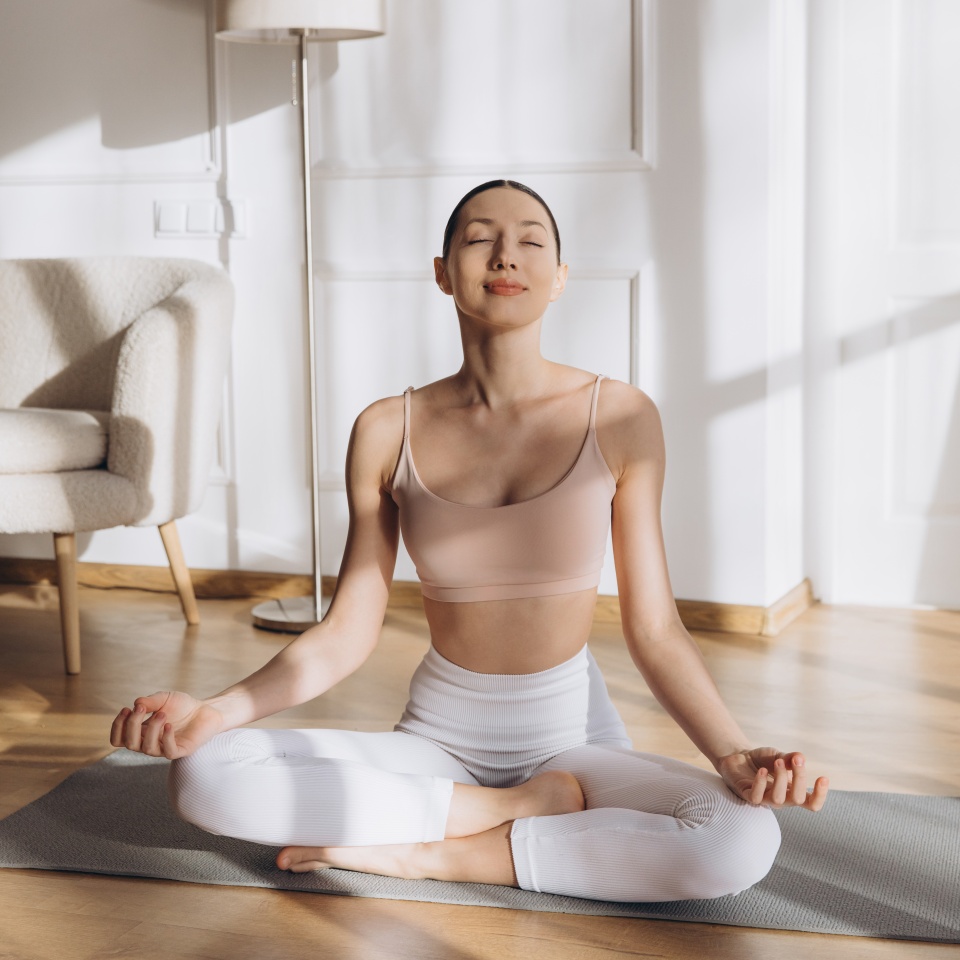 Relaxed woman meditating in lotus pose on yoga mat at home, enjoying peace and mindfulness in her sunlit living room