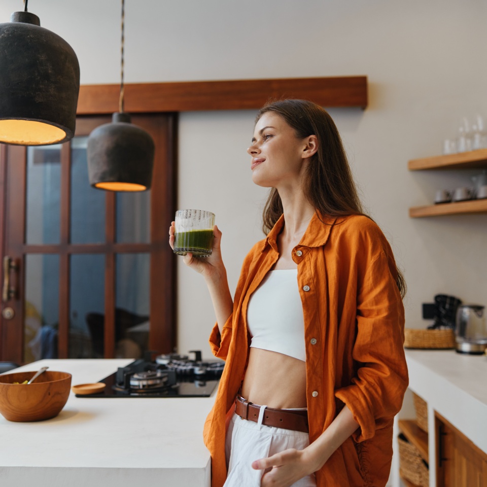 Young woman enjoying a green smoothie in a modern kitchen, wearing an orange shirt and white pants, radiating happiness and wellness in a bright, stylish setting