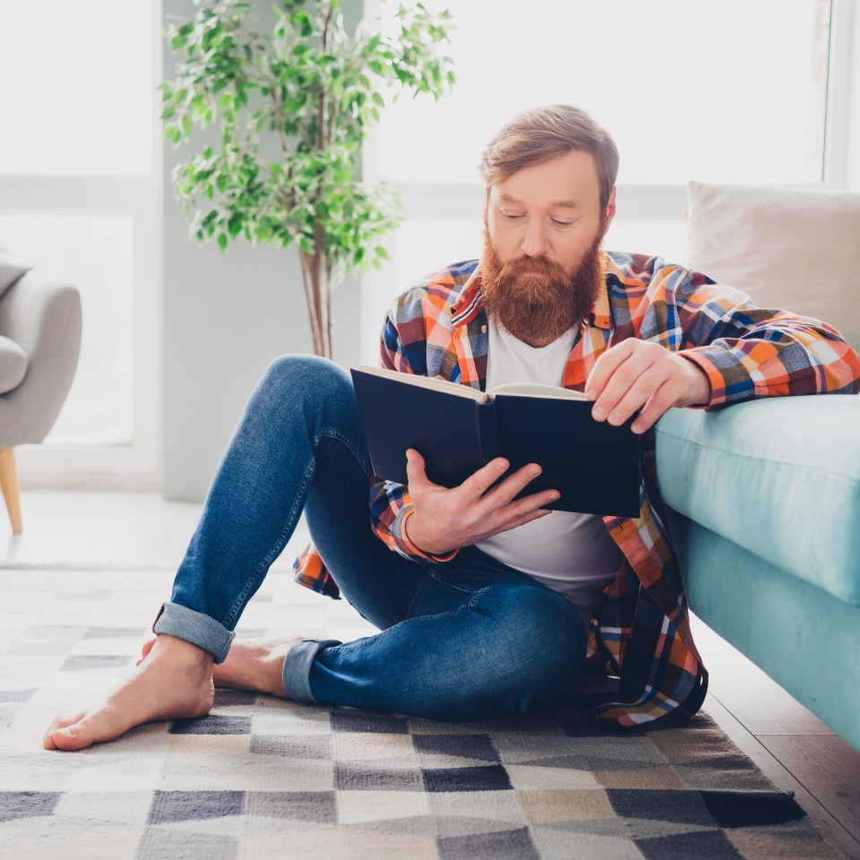 Bearded man reading a book on the floor in a cozy living room with sunlight streaming through the windows