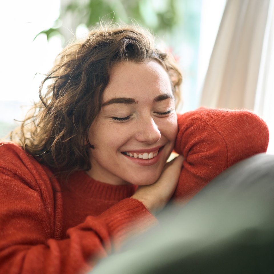 Young smiling pretty curly woman relaxing sitting on couch at home and chilling. Happy relaxed calm beautiful lady enjoying peace of mind and comfort of soft new sofa with eyes closed. Close up.