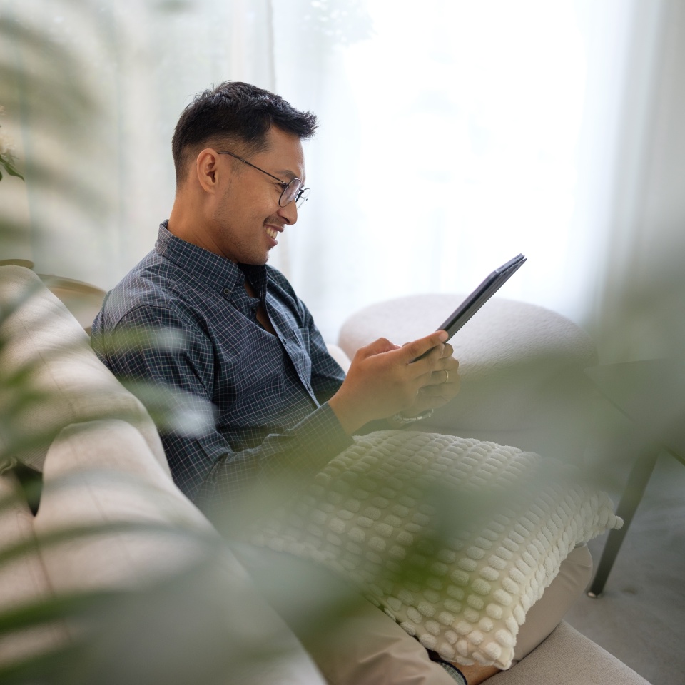 Asian man relaxing on a modern sofa at home, using a digital tablet with a smile, surrounded by indoor plants and natural light in a cozy living room.