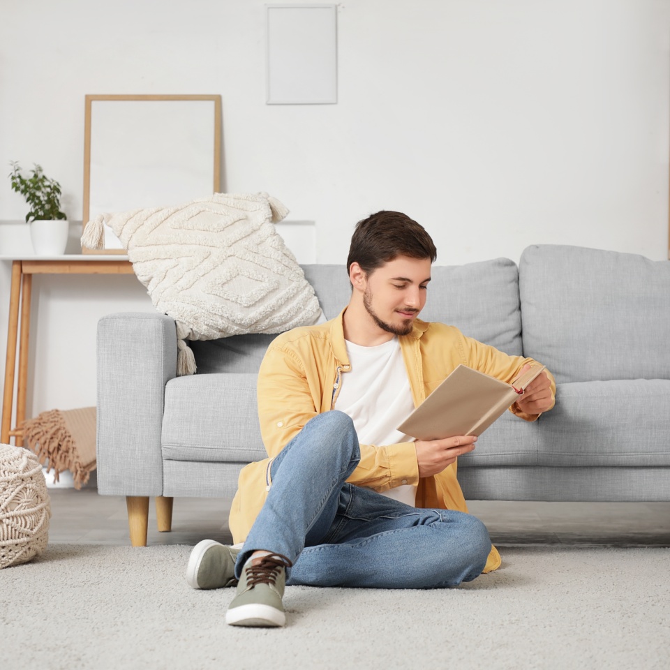 Young man reading book on carpet near sofa at home
