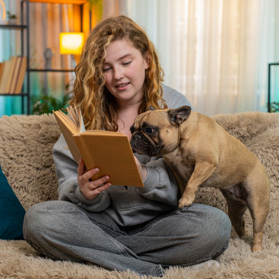 Young relaxed Caucasian redhead woman reads book aloud to pug dog on home sofa with soft voice and warm smile. Pet lies quietly beside her, listening calmly and sharing gentle end-of-day connection.