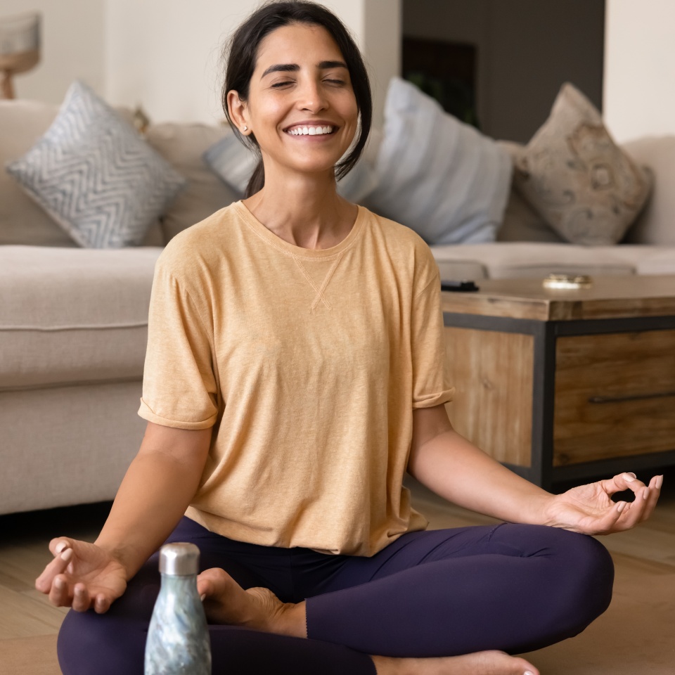 Cheerful Hispanic yogi woman meditating at home with closed eyes, doing yoga on mat unrolled on floor, smiling, laughing, sitting at bottle with cold drink or water. Vertical shot