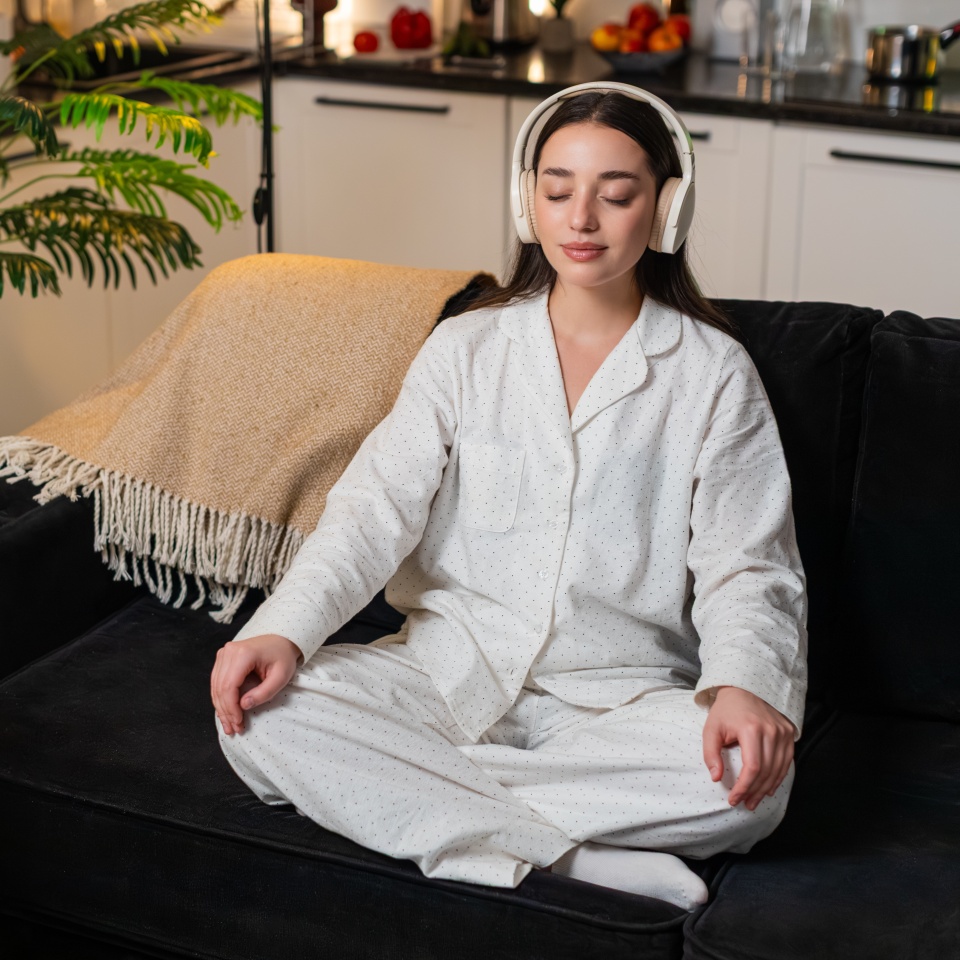 Young woman at night home plays rain track in headphones sits calm breath slow steady finds inner focus. Caucasian girl on sofa at evening eyes closed quiet mood relax chill gentle tone peace in mind.
