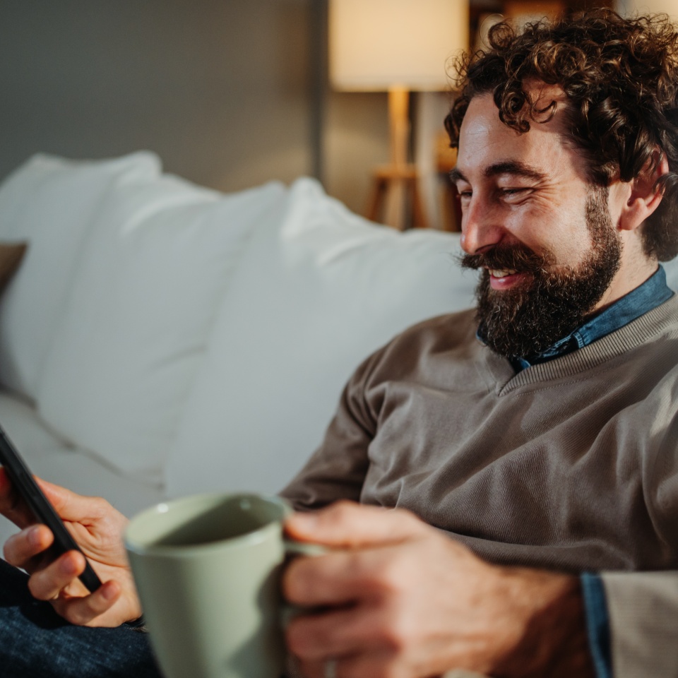 Man relaxing on couch, smiling and using a mobile phone, holding a cup of coffee