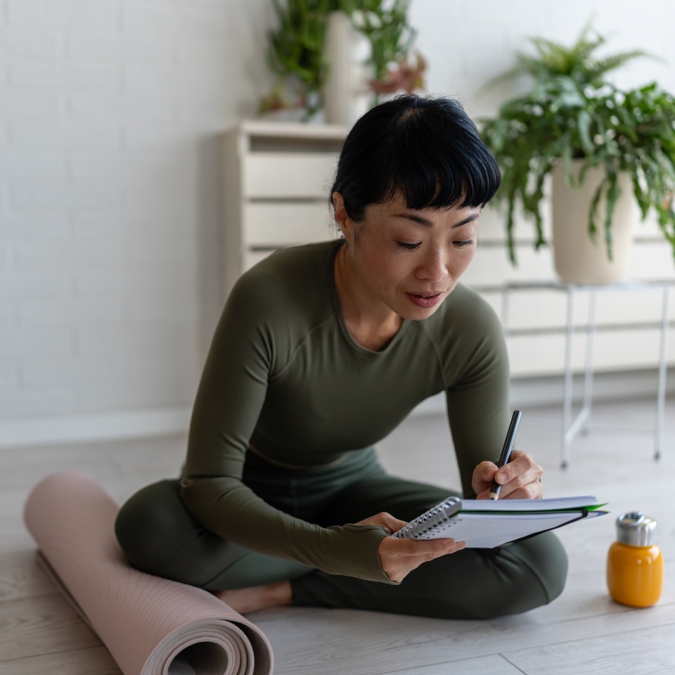 Woman in sportswear sitting on floor by yoga mat, writing in notebook and reflecting after home workout or yoga session