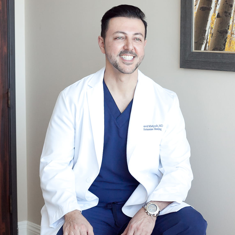 A man wearing a white lab coat over navy scrubs sits on a stool and smiles, with a framed picture on the wall behind him.